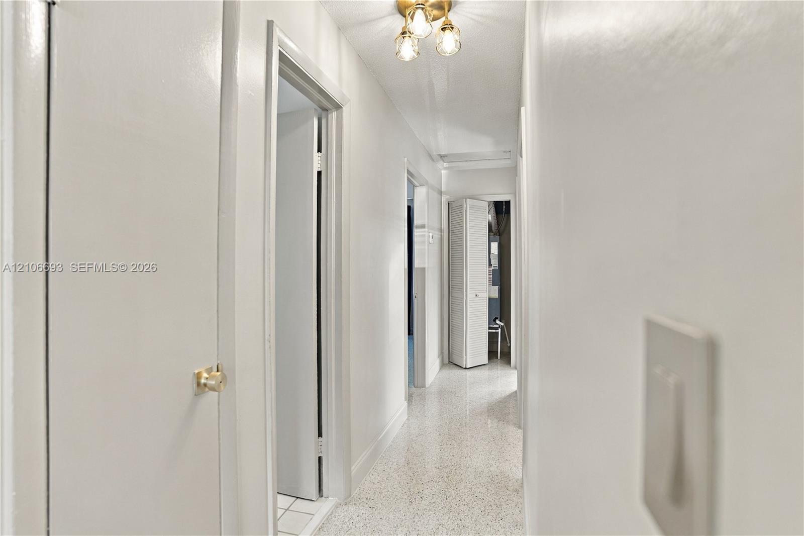 Hallway featuring neutral wall finishes, terrazzo flooring, and a multi-bulb ceiling-mounted light fixture Hallway featuring neutral wall finishes, terrazzo flooring, and a multi-bulb ceiling-mounted light fixture