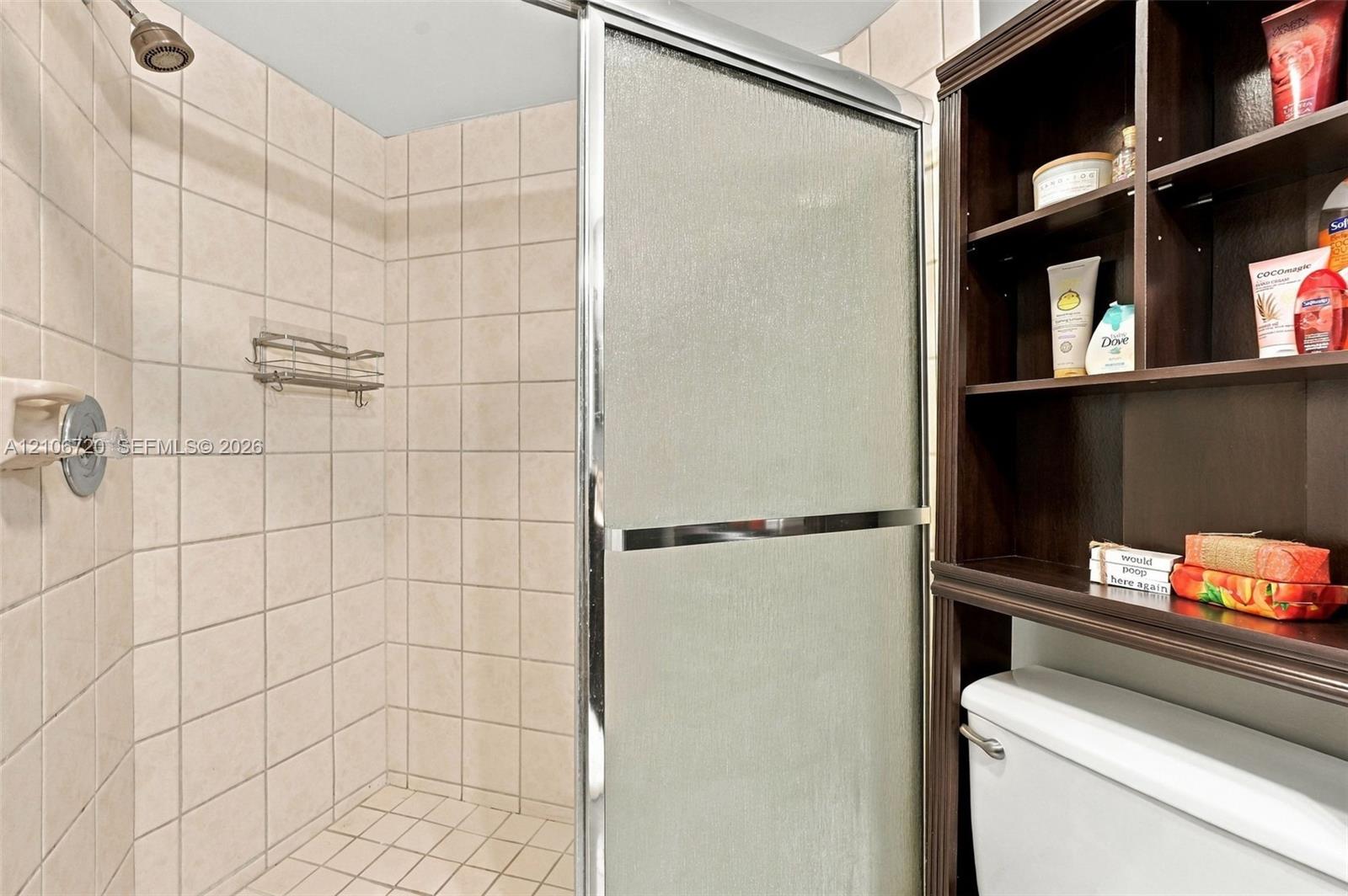 Walk-in shower featuring a sliding glass door, neutral tile surround, wall-mounted caddy, and overhead showerhead, complemented by built-in dark wood shelving above the toilet