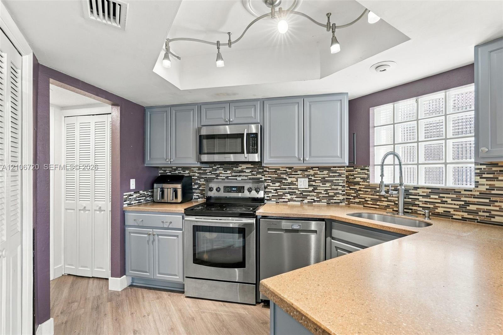 Kitchen featuring shaker-style cabinetry, stainless steel appliances, multi-toned mosaic tile backsplash, solid-surface countertops, and a glass block window
