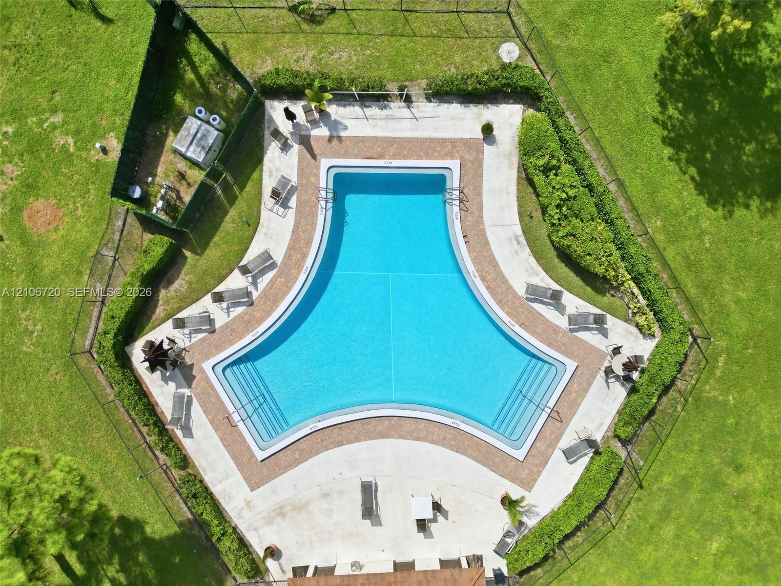 T-shaped pool with integrated stairs, surrounded by a paved deck, enclosed by perimeter fencing and landscaped hedges within a grass lawn