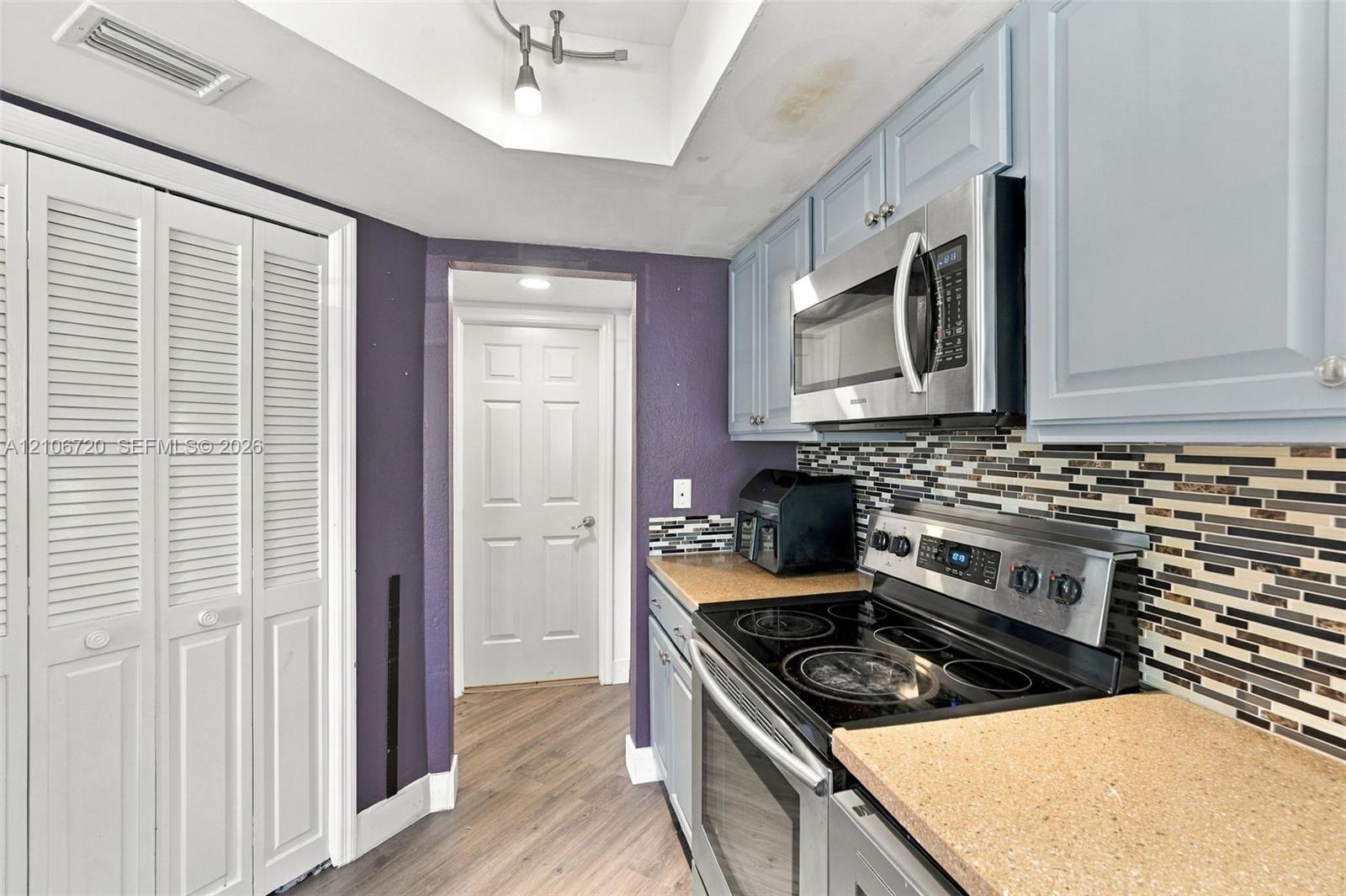 Kitchen featuring light blue cabinetry, stainless steel microwave, glass-top electric range, multi-toned mosaic tile backsplash, and wood-finish flooring