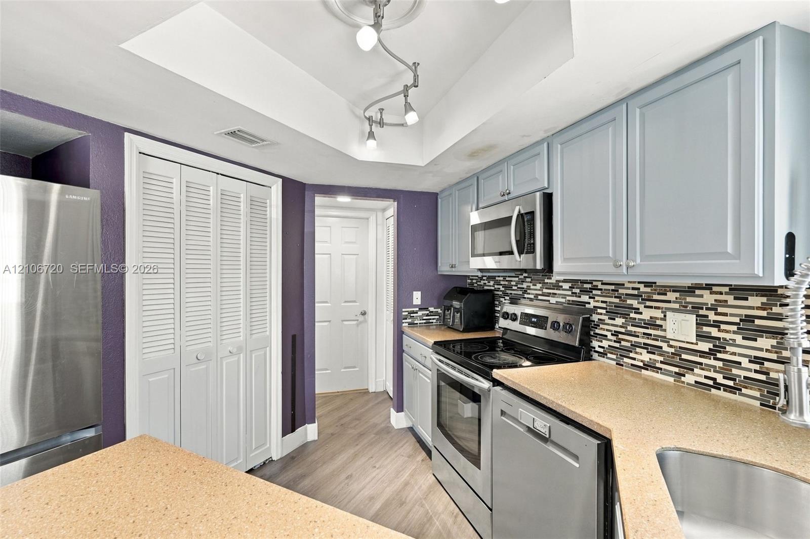 Kitchen featuring stainless steel appliances, multi-toned tile backsplash, solid-surface countertops, light-finish cabinetry, and wood-finish flooring