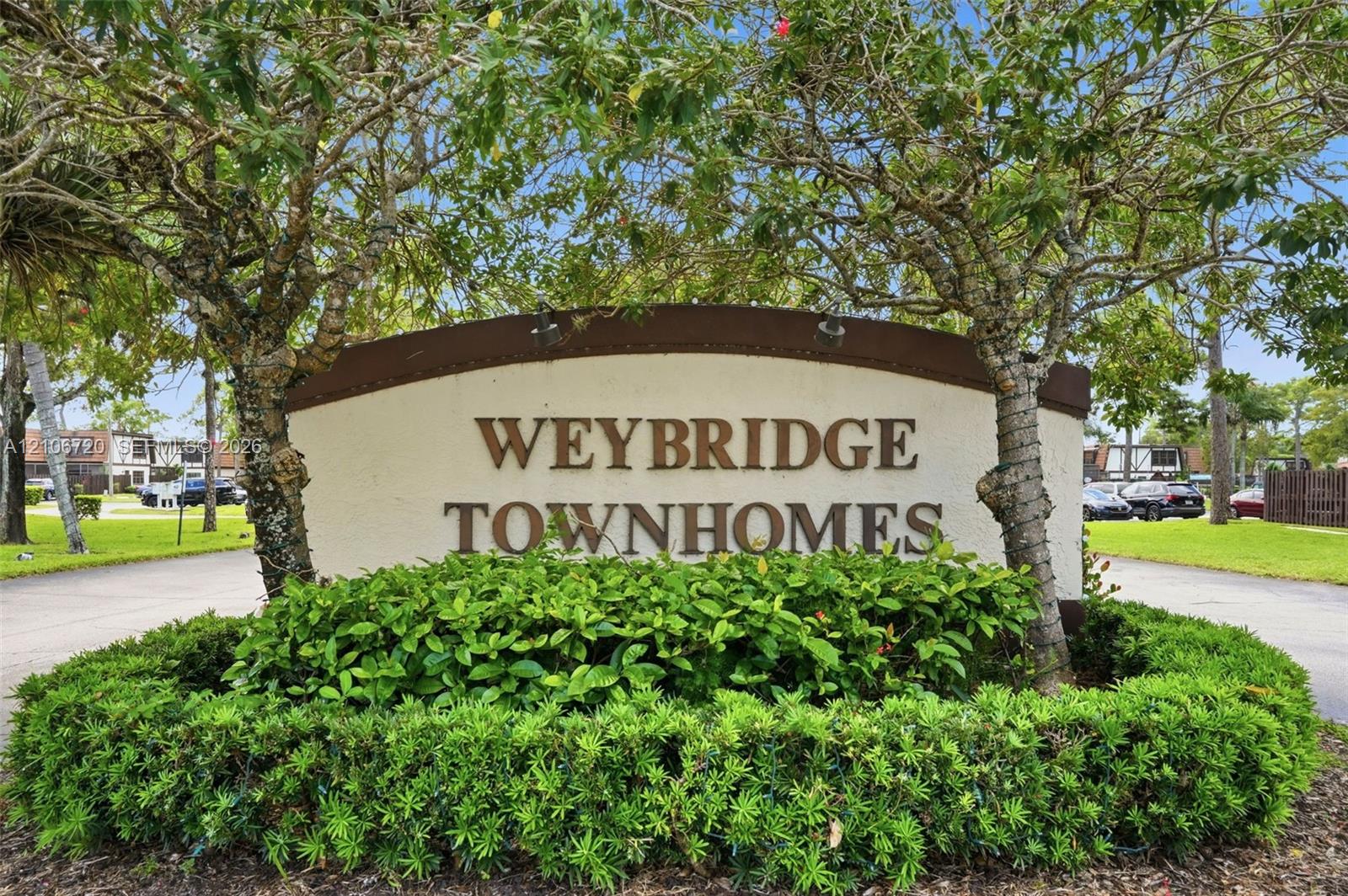 Weybridge Townhomes monument sign featuring dimensional lettering, decorative arch-top trim, and integrated landscape lighting, surrounded by manicured shrubbery and mature canopy trees