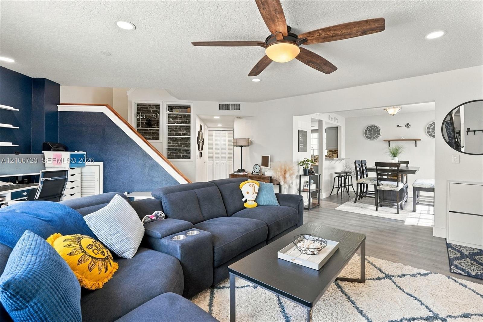 Open-concept living space featuring wood-finish flooring, recessed lighting, a ceiling fan with integrated lighting, and an accent wall with exposed brick detailing