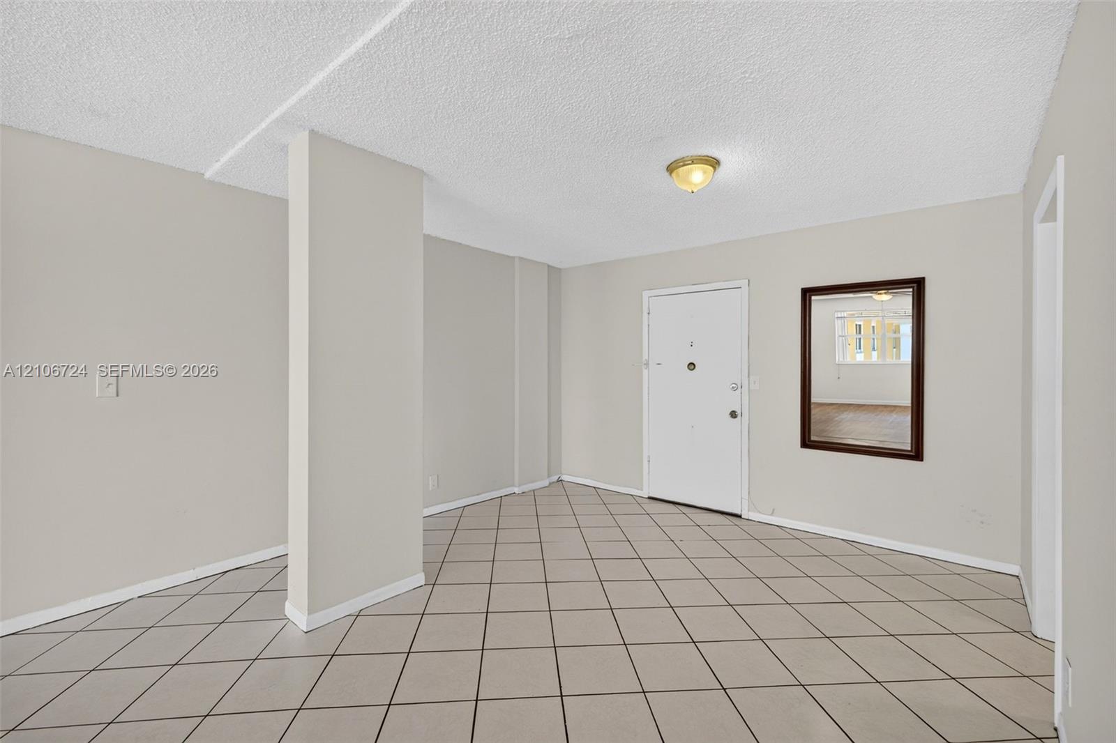 Neutral-toned interior space featuring ceramic tile flooring, a textured ceiling, a flush-mount light fixture, and a wall-mounted mirror