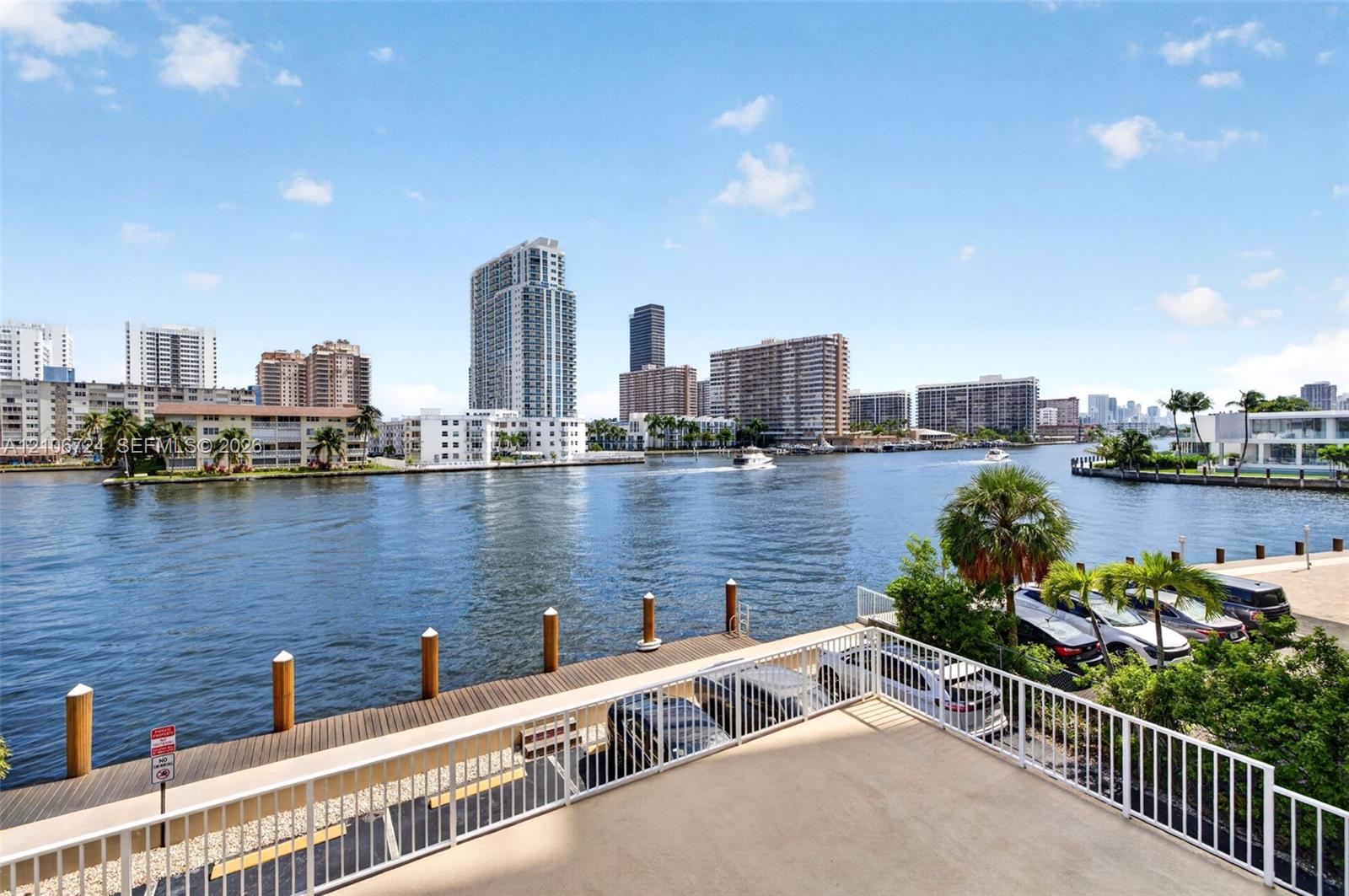 Concrete balcony with white metal railings, overlooking a waterway, wooden dock, and urban skyline