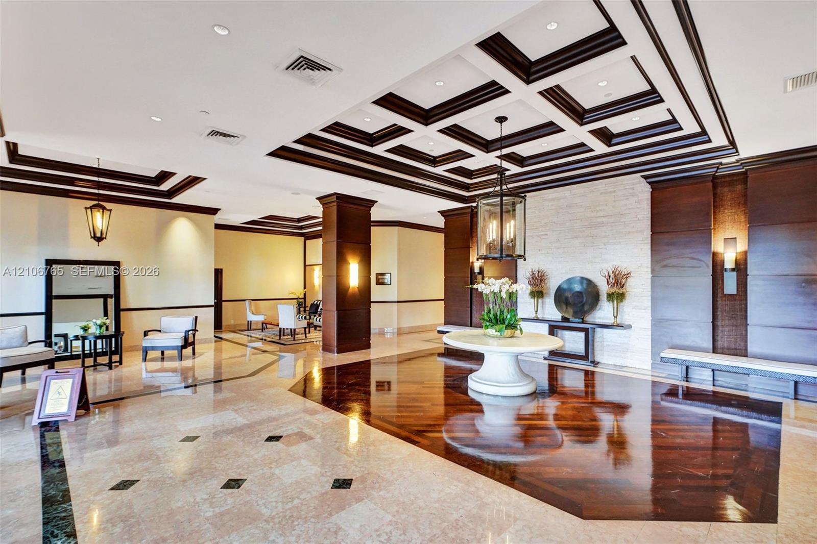 Coffered ceiling with dark wood detailing, inlaid stone flooring, textured stone accent wall, wood-paneled columns, and lantern-style light fixtures.