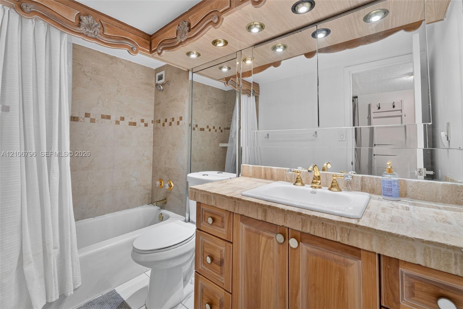 Bathroom featuring a vanity with a stone countertop, inset sink, brass-finish faucet, custom wood cabinetry, and recessed lighting integrated into a decorative wood-trimmed mirror surround, with stone-tile tub surround and decorative mosaic inlay
