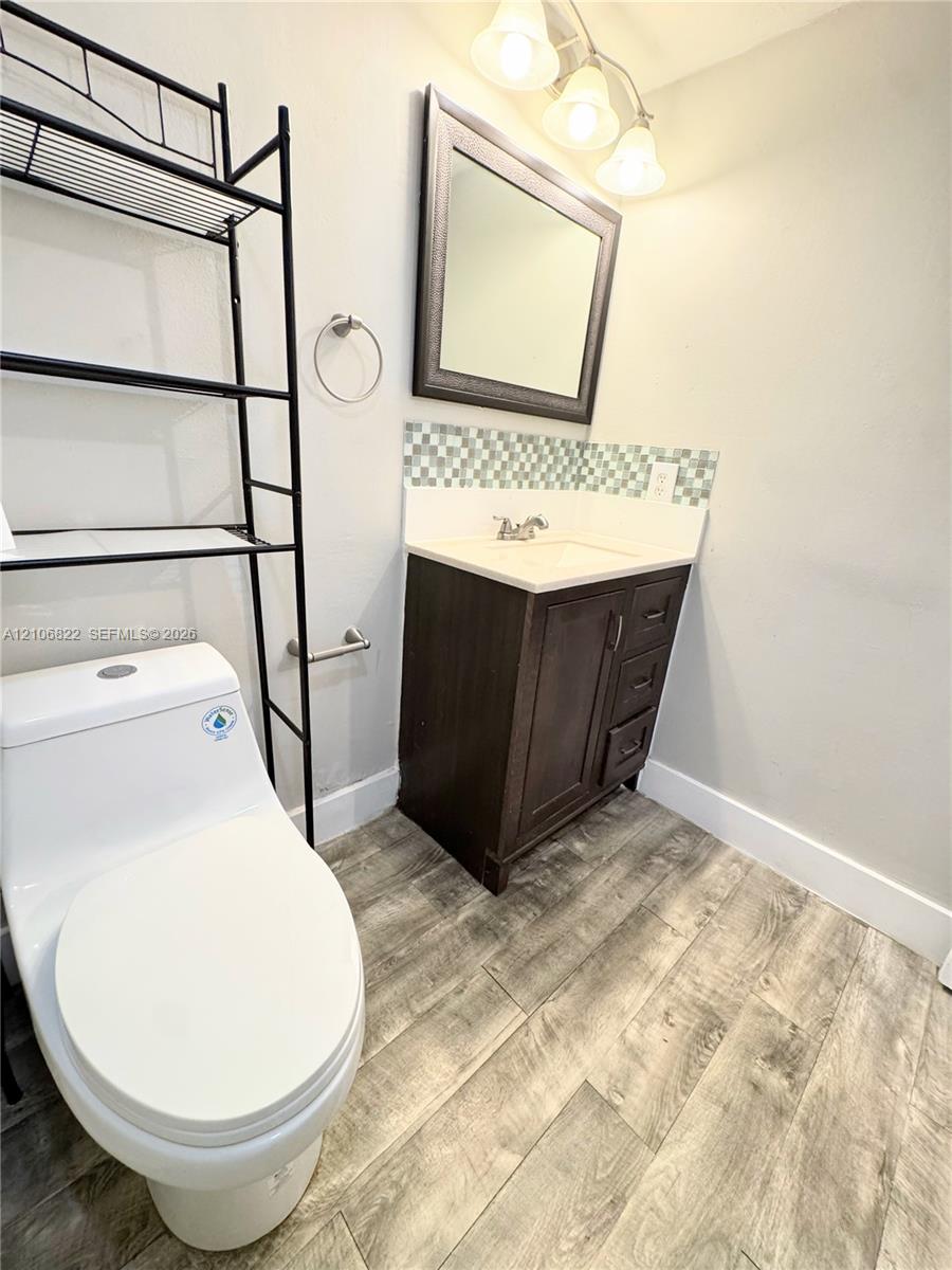 Dark-finish vanity with a rectangular sink, patterned glass mosaic backsplash, wood-finish flooring, and a framed mirror