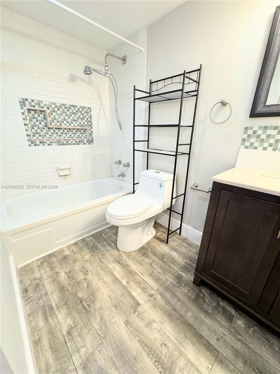 Bathroom featuring wood-finish flooring, a tub-shower combination with white wall tile, a tiled mosaic inlay, a dark wood-finish vanity, and a dual-handle shower faucet