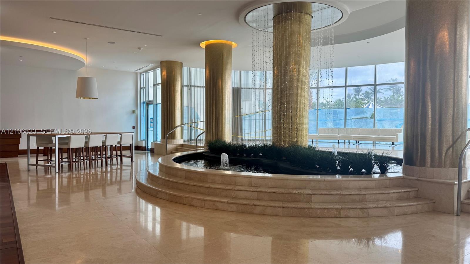 Grand lobby featuring polished stone flooring, circular tiered water feature, gold-tiled support columns, full-height floor-to-ceiling windows, and recessed cove lighting