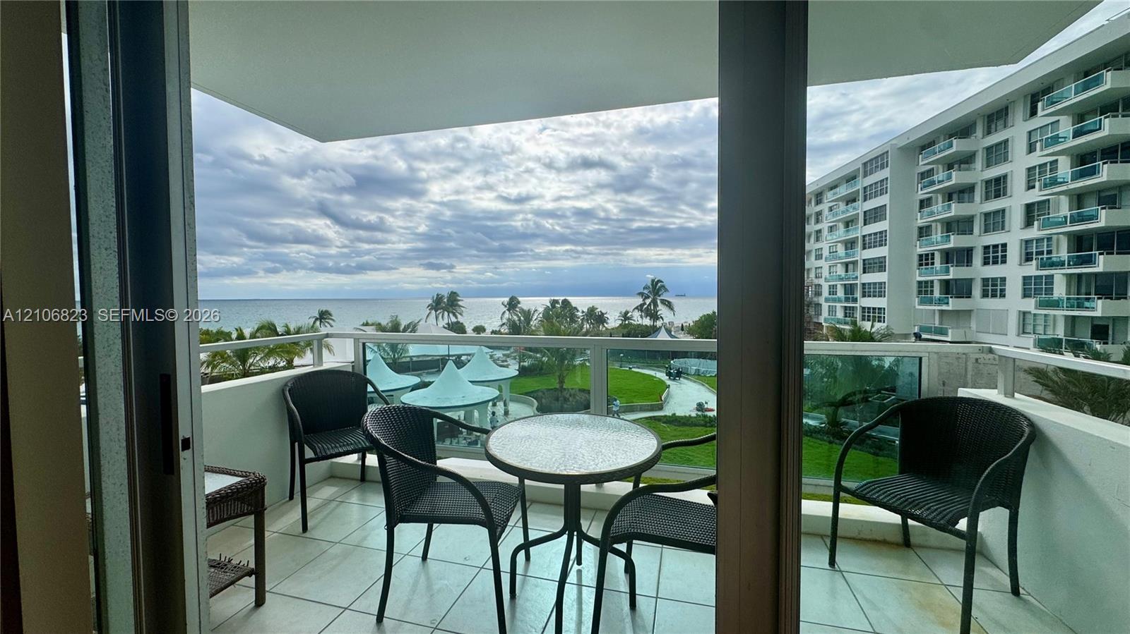 Covered balcony featuring tile flooring and a glass-paneled railing overlooking the ocean and landscaped grounds