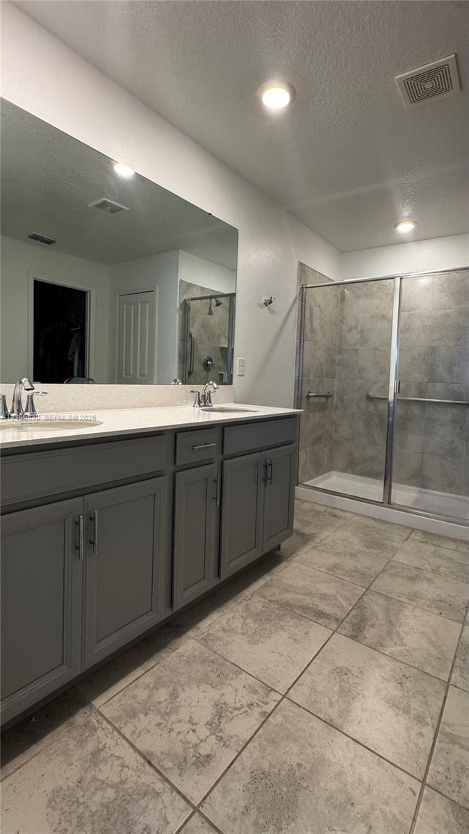 Dual-sink vanity with gray shaker-style cabinetry, white countertop, large wall-to-wall mirror, and glass-enclosed shower with stone-finish tile surround
