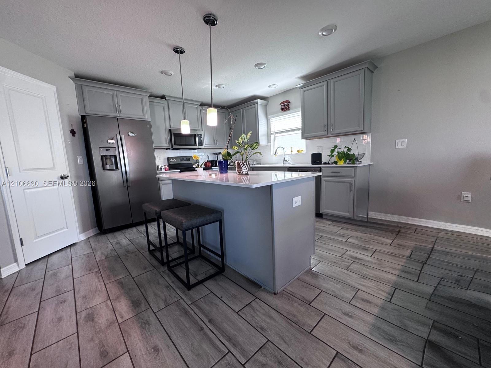 Kitchen featuring gray shaker-style cabinetry, central island with stone-finish countertop, stainless steel refrigerator, recessed lighting, and wood-finish tile flooring Kitchen featuring gray shaker-style cabinetry, central island with stone-finish countertop, stainless steel refrigerator, recessed lighting, and wood-finish tile flooring