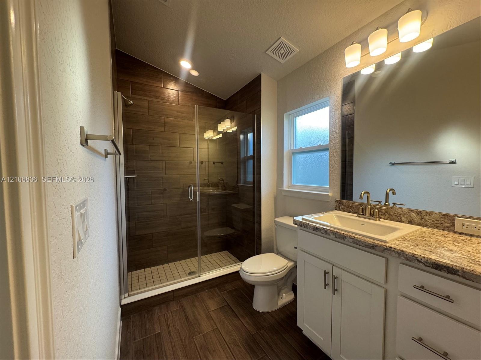 Bathroom featuring a walk-in shower with glass enclosure, wood-finish tile wall surround, stone-composite countertop, white vanity cabinetry with brushed-metal hardware, and wood-finish flooring