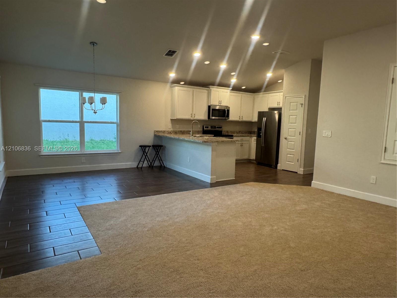 Open-concept living space featuring wood-finish tile flooring, neutral carpeting, white shaker-style cabinetry, a breakfast bar with stone-finish countertops, and stainless steel appliances Open-concept living space featuring wood-finish tile flooring, neutral carpeting, white shaker-style cabinetry, a breakfast bar with stone-finish countertops, and stainless steel appliances