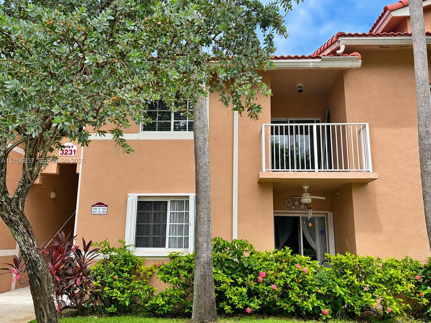 Stucco exterior featuring a terracotta-tiled roof, second-story balcony with metal railings, and established perimeter landscaping