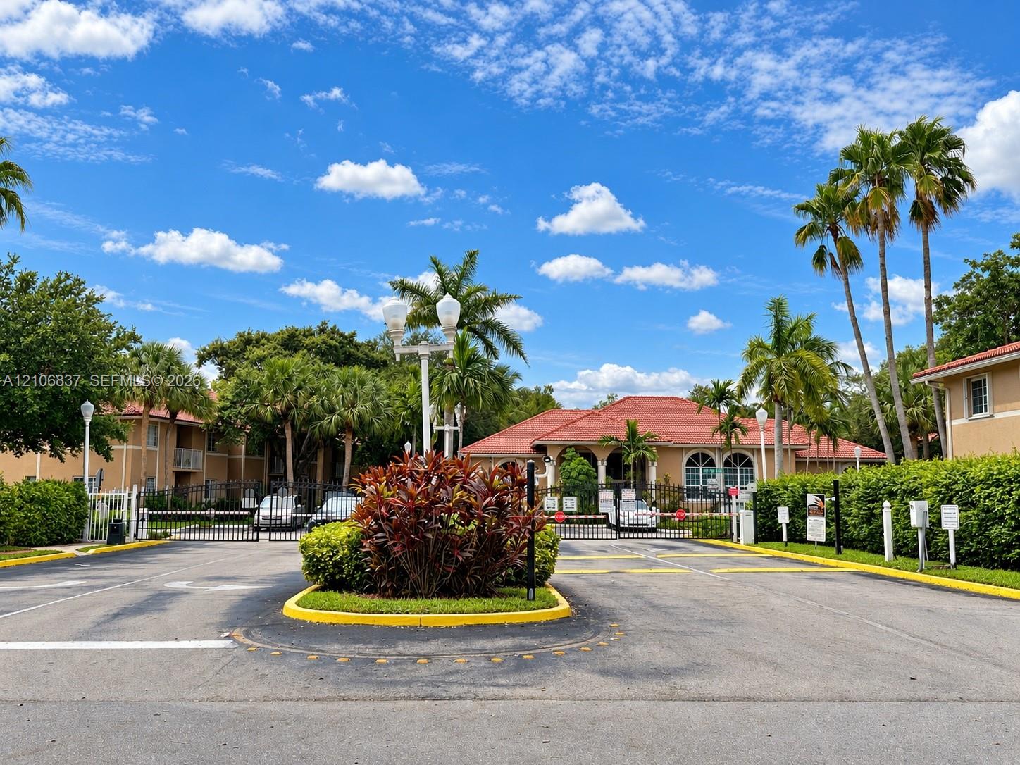 Gated entrance featuring a paved arrival court, perimeter fencing, mature palm landscaping, and tile roof architecture