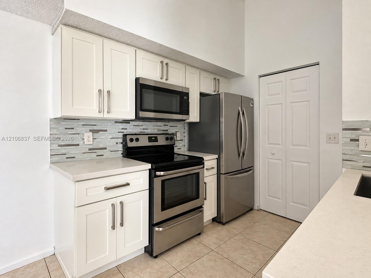 Kitchen featuring white shaker-style cabinetry, quartz countertops, multi-toned linear tile backsplash, stainless steel appliances, and tile flooring