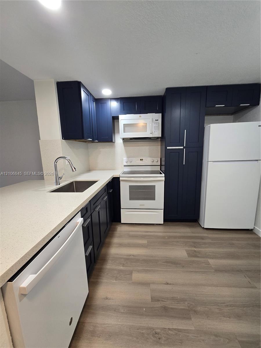 Kitchen featuring navy cabinetry, solid-surface countertops, wood-finish flooring, stainless steel sink with high-arch faucet, and white appliances