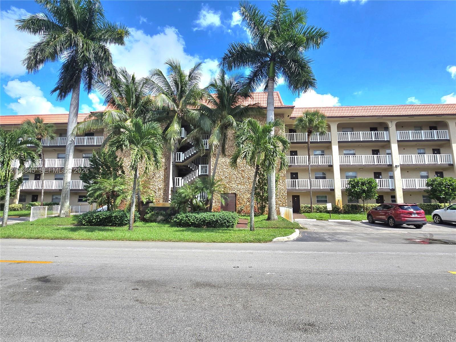 Multi-story building exterior featuring stone masonry cladding, a terracotta tile roof, and wrap-around balconies with white railings, set amidst mature palm trees and landscaped grounds