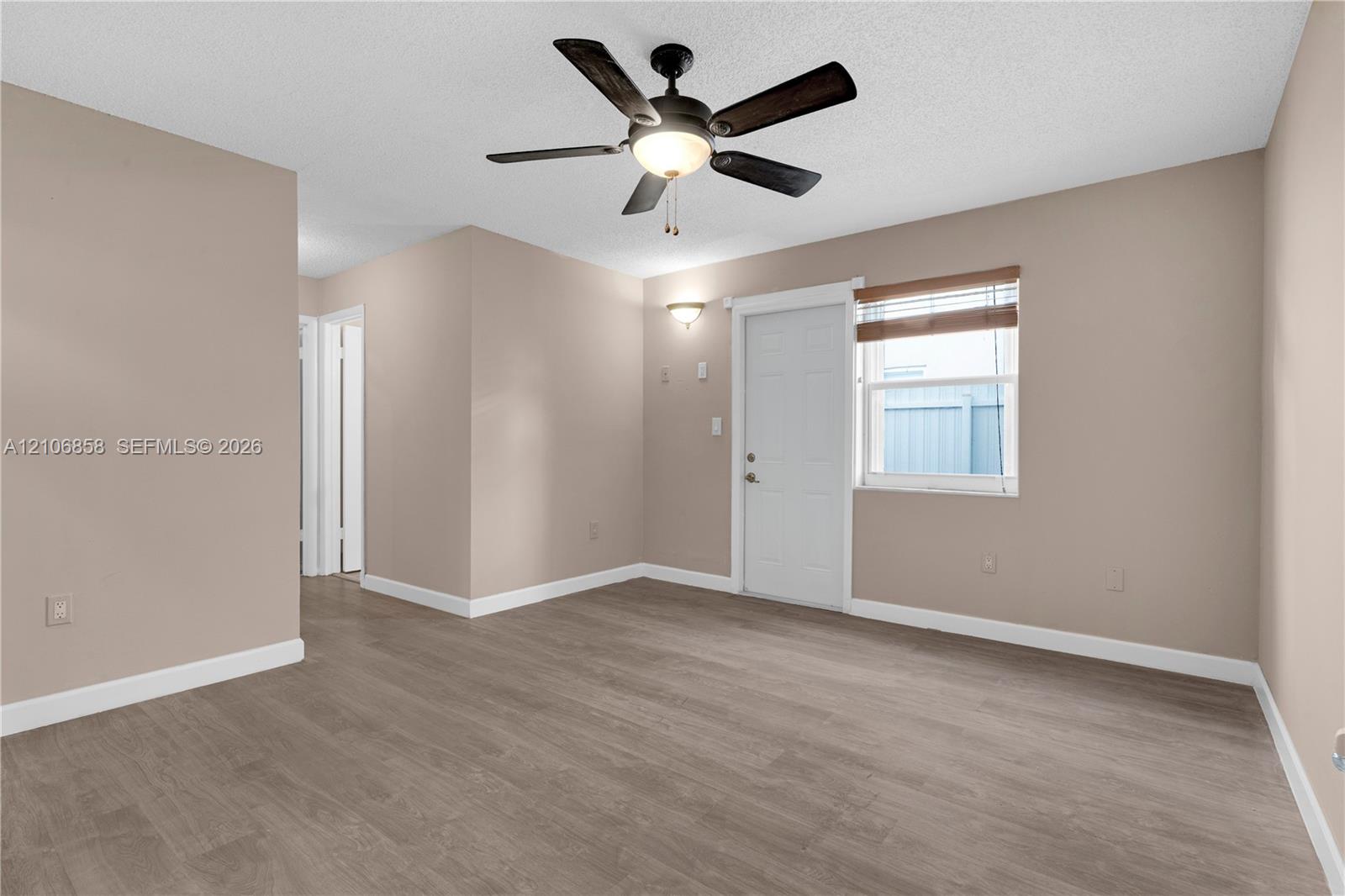 Neutral-toned room featuring wood-finish flooring, a central ceiling fan with integrated lighting, wall-mounted accent lighting, and a single-panel door