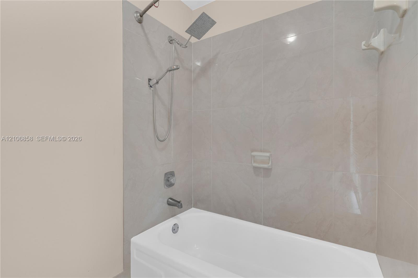 Bathtub surround with neutral-toned oversized wall tile, rainfall shower head, handheld shower fixture, and integrated soap niche