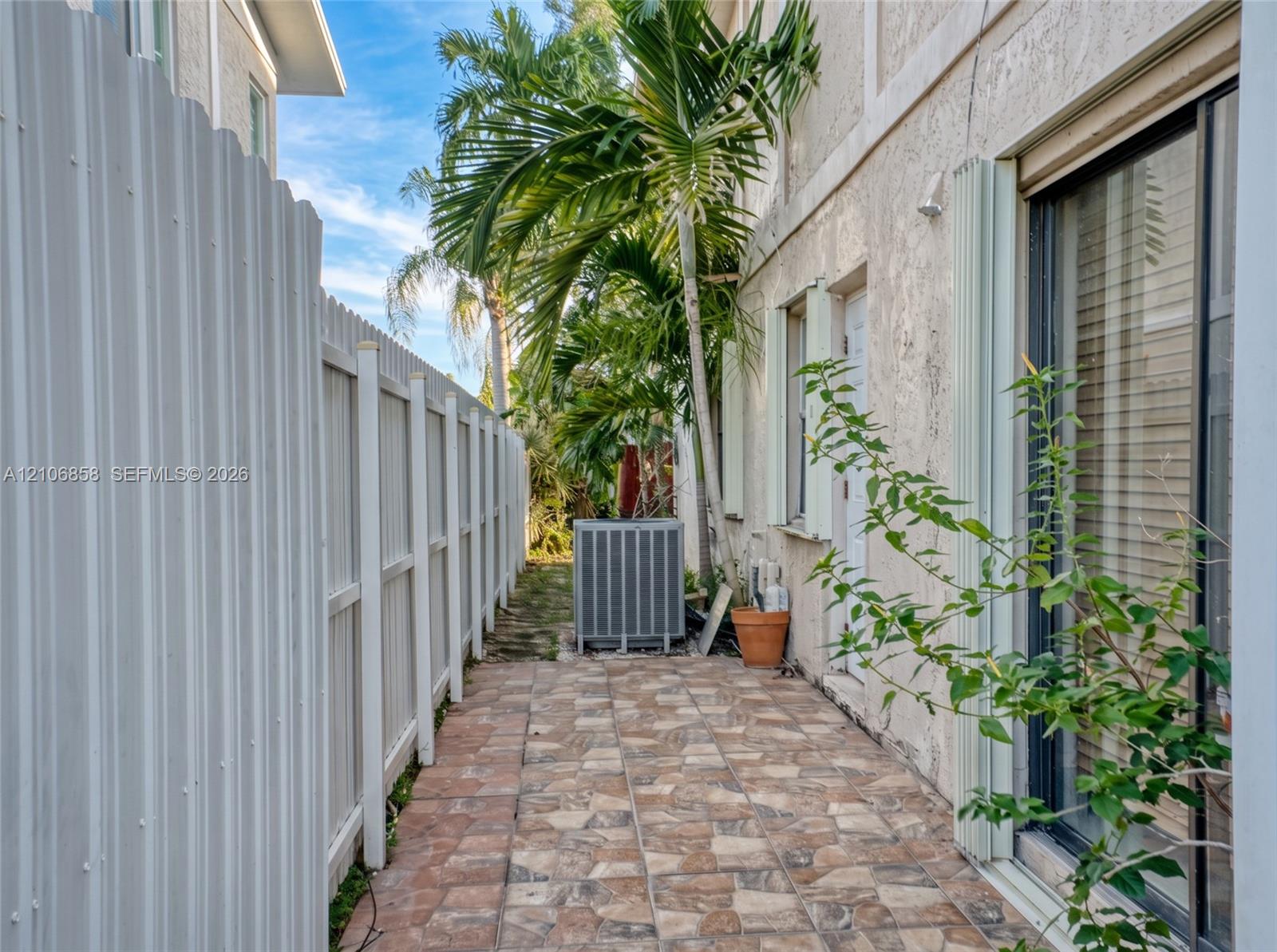 Side exterior featuring a paved stone-patterned walkway, white corrugated metal fencing, and stucco siding