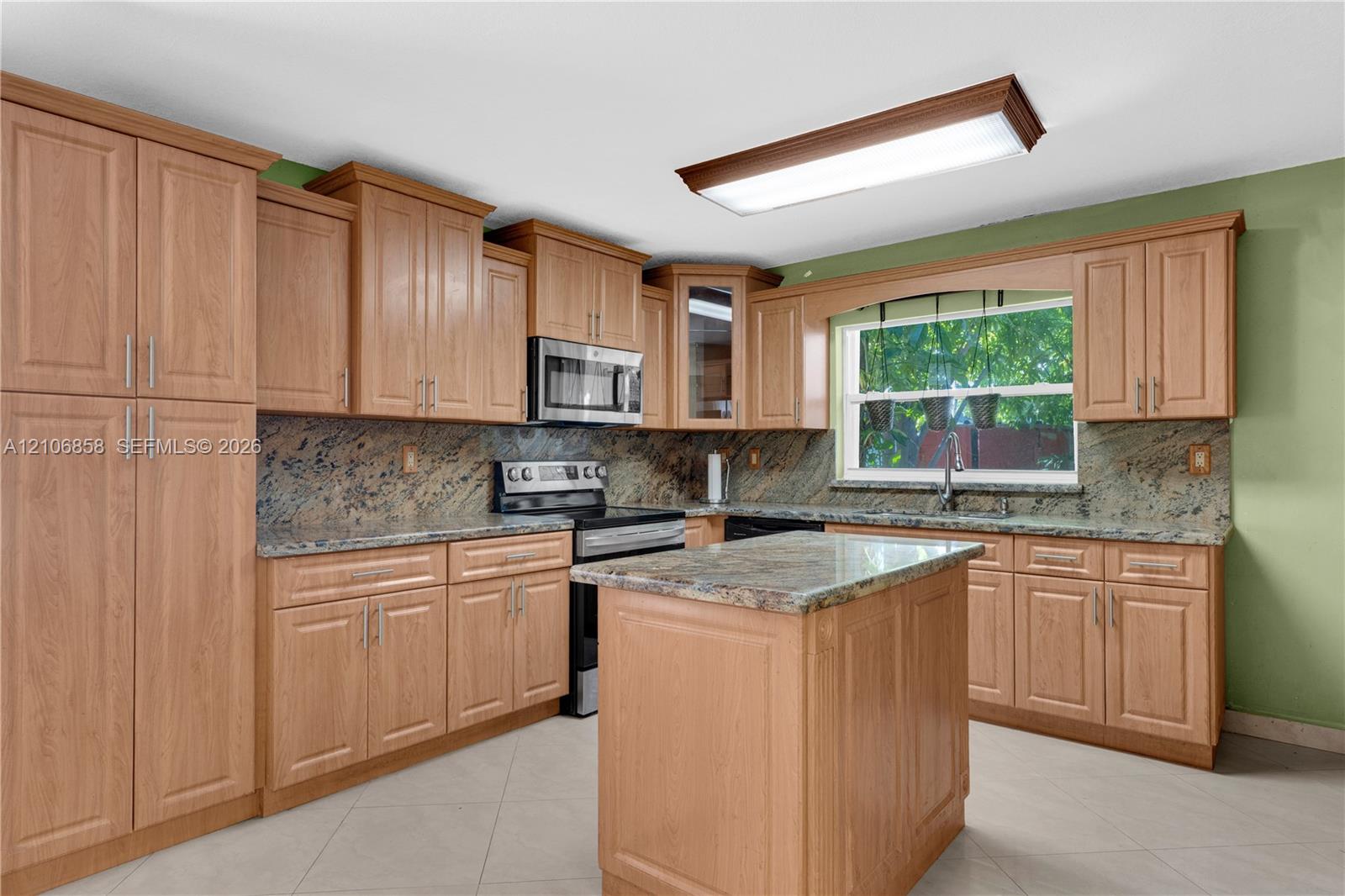 Kitchen featuring wood-finish cabinetry, natural stone countertops, a center island, stainless steel microwave, and tile flooring Kitchen featuring wood-finish cabinetry, natural stone countertops, a center island, stainless steel microwave, and tile flooring