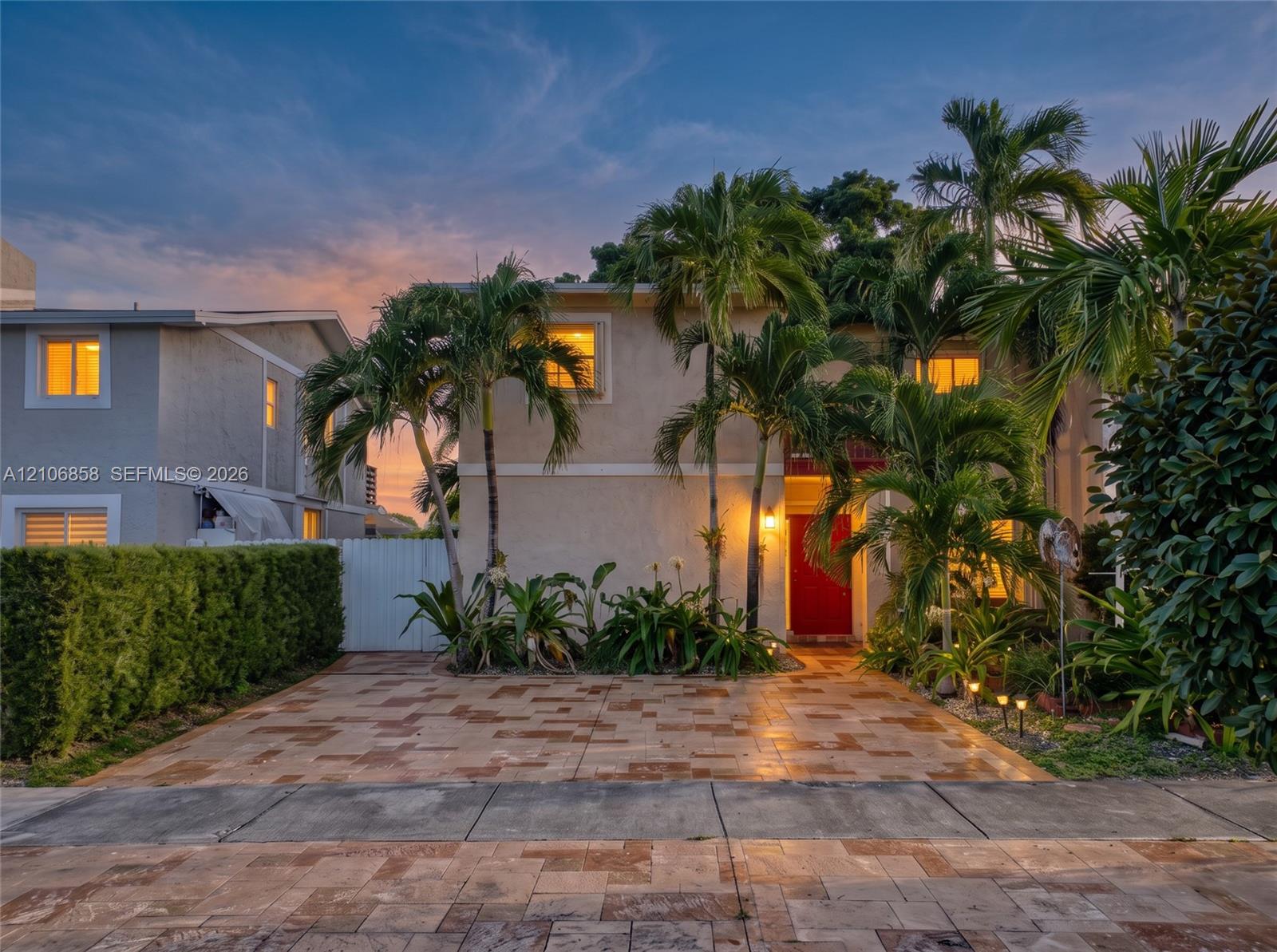 Two-story stucco exterior featuring a paved stone driveway, mature palm trees, manicured hedges, and a vibrant red front door