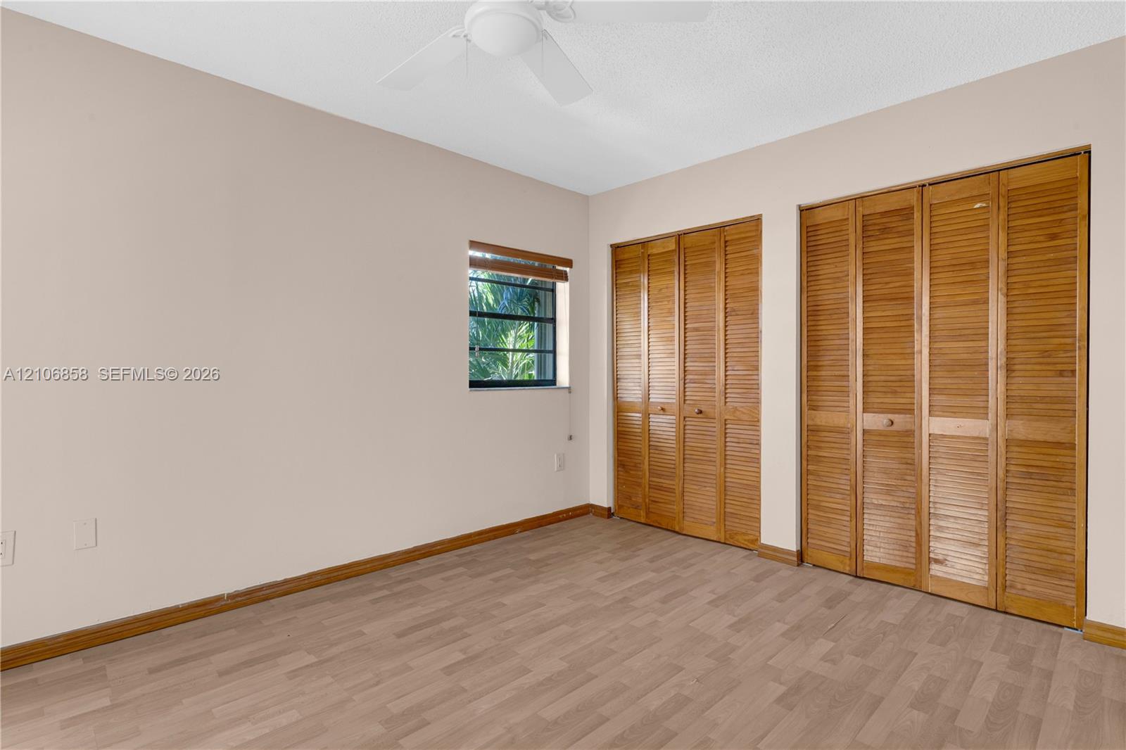 Interior space featuring wood-finish flooring, two sets of bifold wooden closet doors, a ceiling fan with integrated lighting, and a window with wood-slat blinds