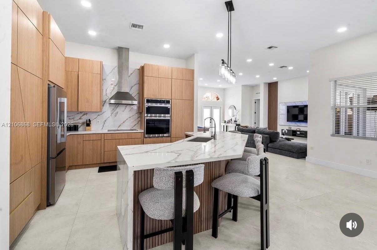Kitchen featuring flat-panel wood-finish cabinetry, stone-finish waterfall edge island, stainless steel wall-mounted oven and microwave, stone-finish backsplash, and recessed ceiling lighting