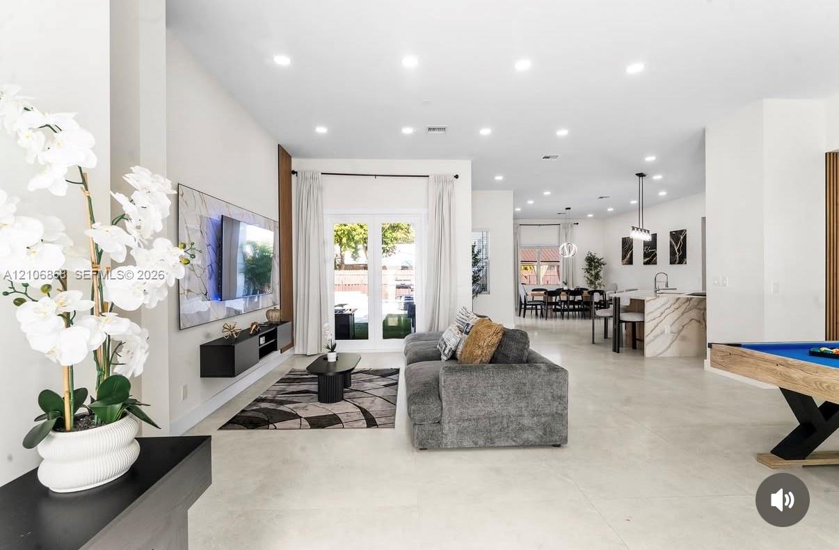 Open-concept interior featuring large-format tile flooring, recessed ceiling lighting, a wall-mounted floating media console, and a kitchen island with a waterfall-edge countertop