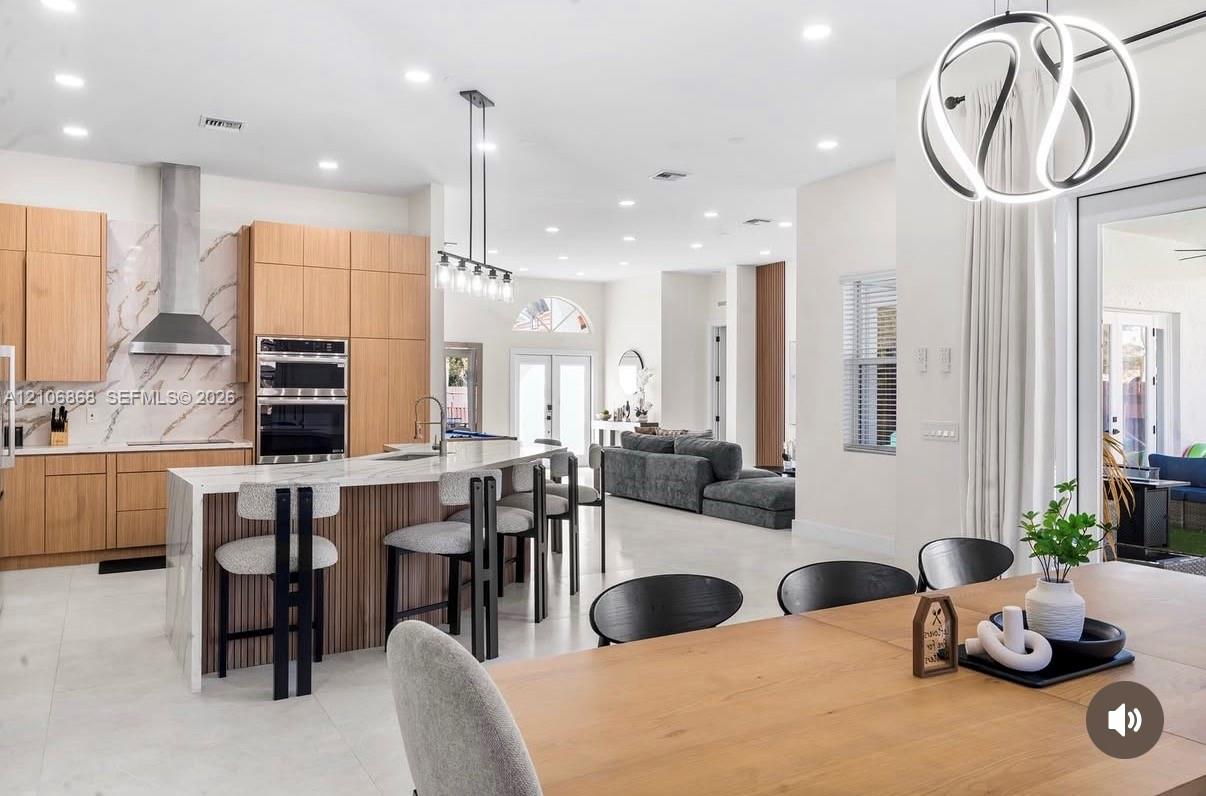 Kitchen featuring light wood-finish cabinetry, stone-style countertops with a waterfall edge, stainless steel built-in appliances, recessed lighting, and a modern sculptural pendant fixture