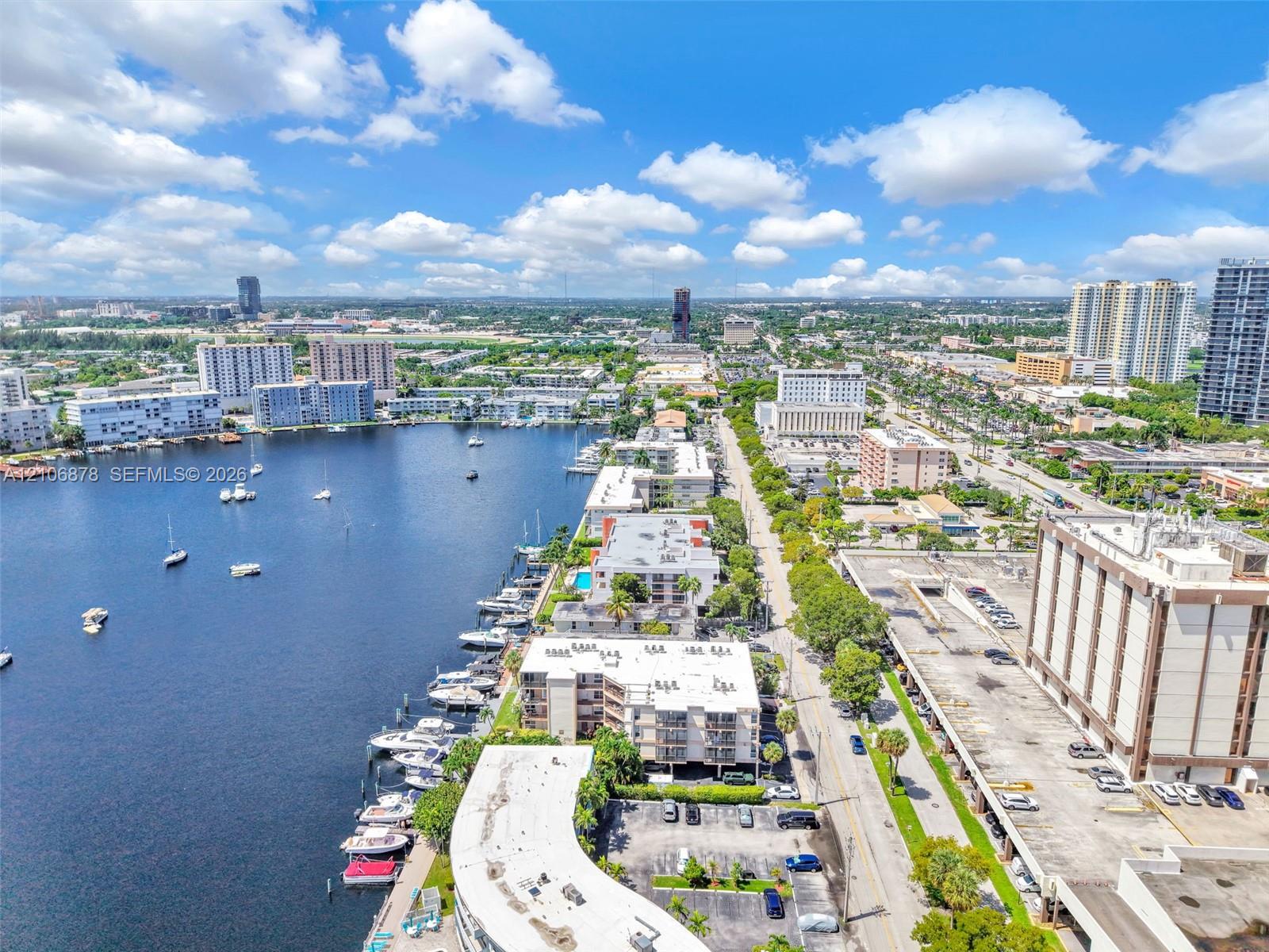 Aerial perspective of a waterfront setting featuring a canal, multi-level residential structures, surface parking, and urban infrastructure