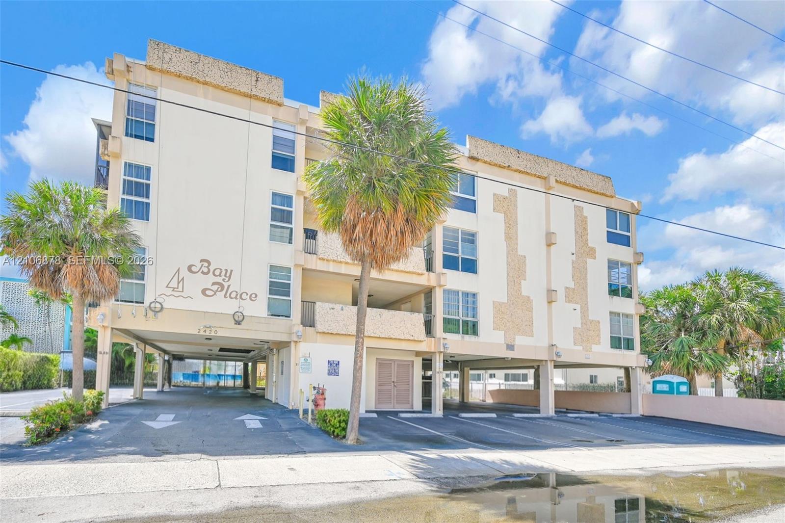 Multi-story residential building exterior featuring a stucco facade, textured stone accents, ground-level covered parking, and mature palm trees