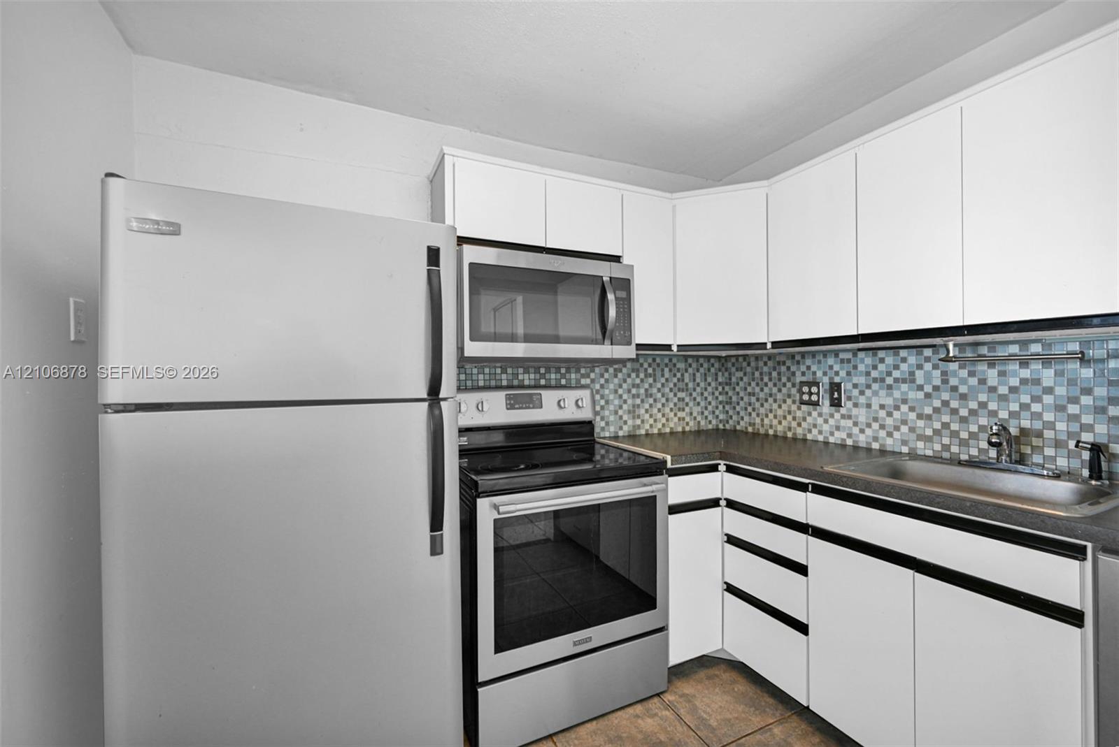 Kitchen featuring white cabinetry, stainless steel appliances, multi-toned mosaic tile backsplash, dark countertop, and tiled flooring