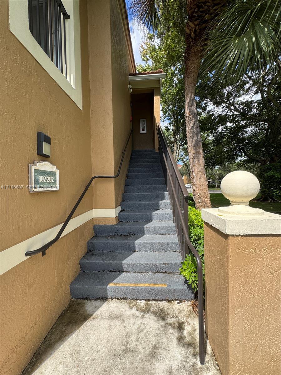 Stucco exterior featuring concrete stairs, a metal handrail, a decorative stone-finish address plaque, and a masonry pillar topped with a spherical finial