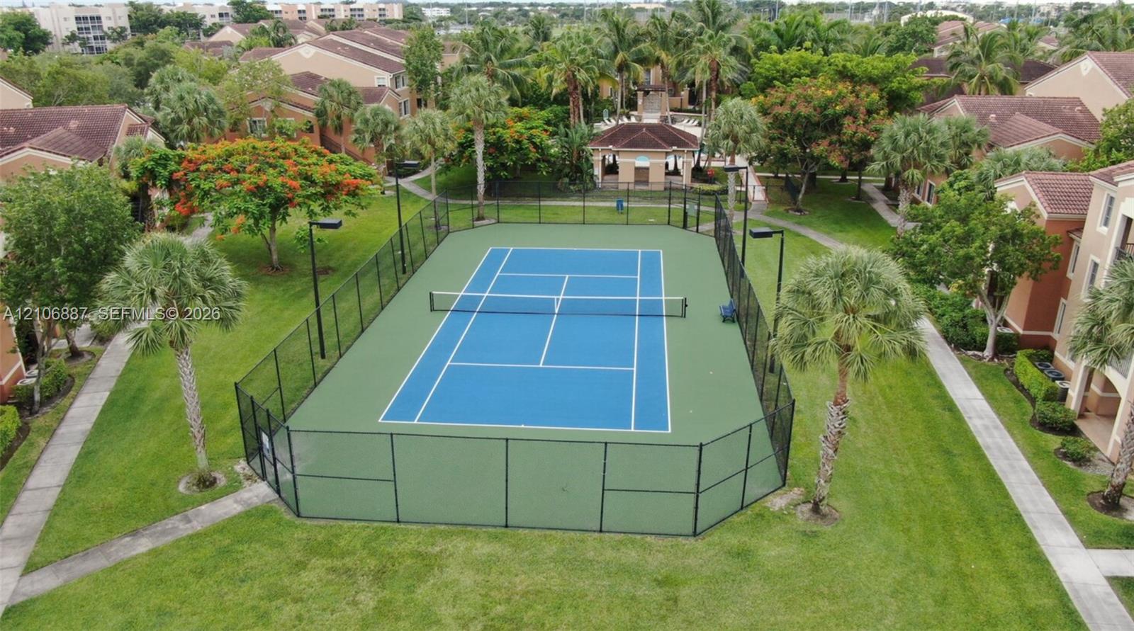 Tennis court featuring a paved surface, perimeter fencing, high-mast lighting, and surrounding landscaped grounds