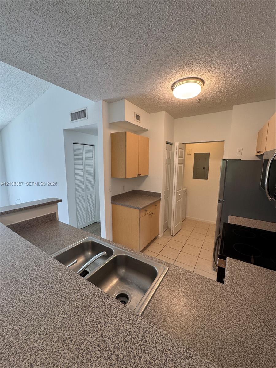 Kitchen featuring a dual-basin stainless steel sink, solid-surface countertops, light wood-finish cabinetry, and ceramic tile flooring
