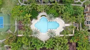 Aerial perspective of a common area featuring a swimming pool, paved deck, hard-surface tennis court, and lush landscaping with mature palm trees