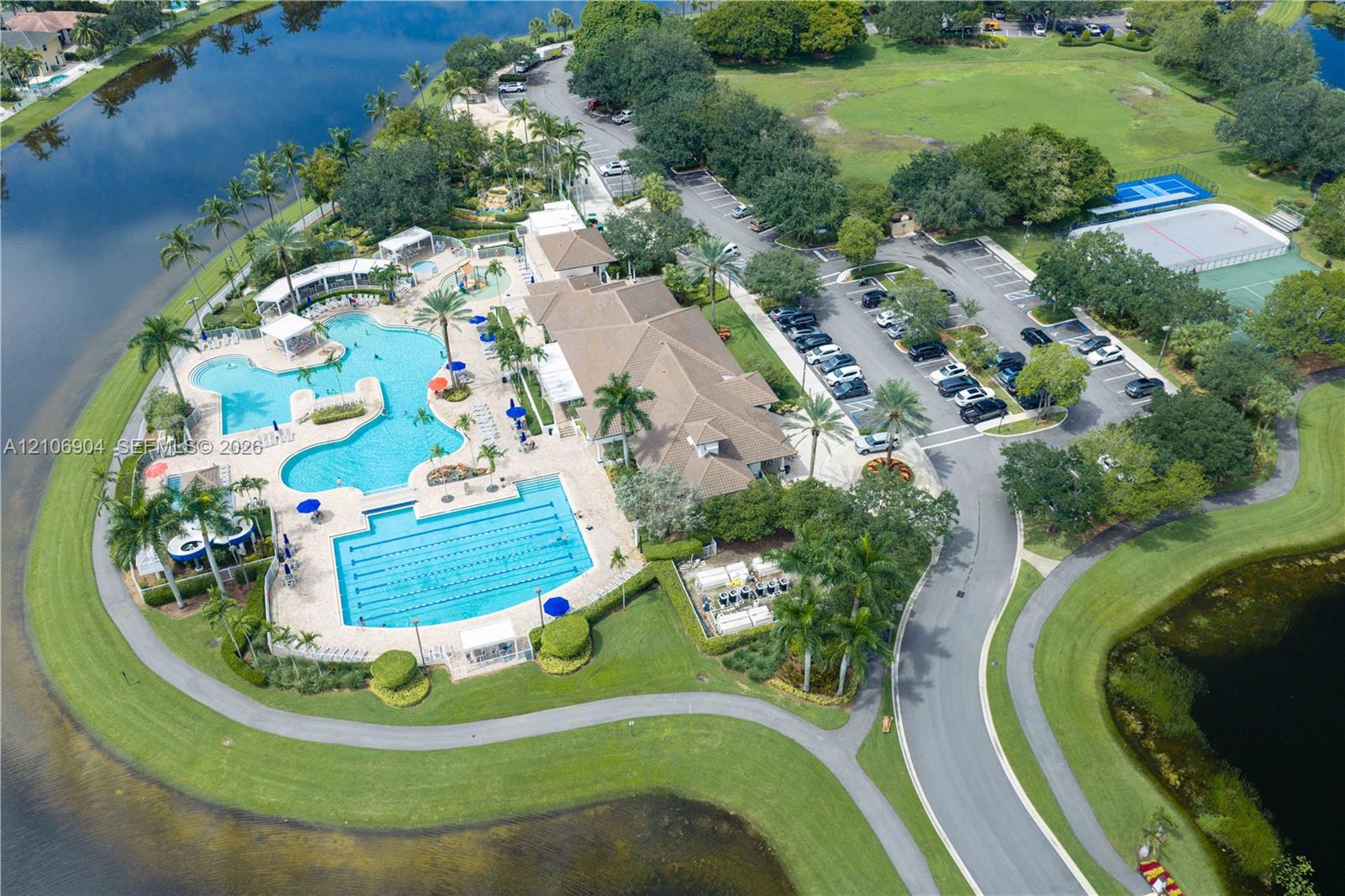 Recreational complex featuring multiple swimming pools, paved walking paths, tennis courts, and landscaped grounds situated along a water feature