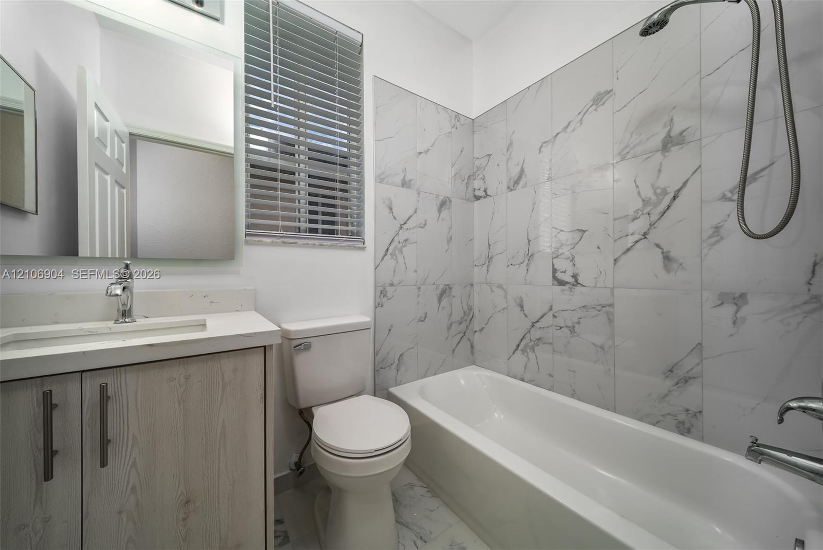 Bathroom featuring a shower-tub combination with white and gray veined tile surround, vanity with stone-finish countertop, undermount sink, and wall-mounted mirror