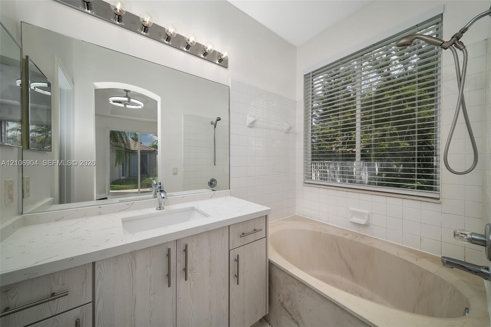 Bathroom featuring a light-toned wood-finish vanity with a rectangular sink, quartz countertop, large wall-to-wall mirror, oval soaking tub, and subway tile wall surround