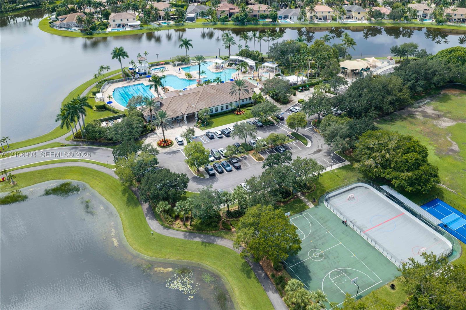 Aerial perspective of community facilities featuring a swimming pool, basketball court, tennis court, and waterfront grounds with mature landscaping