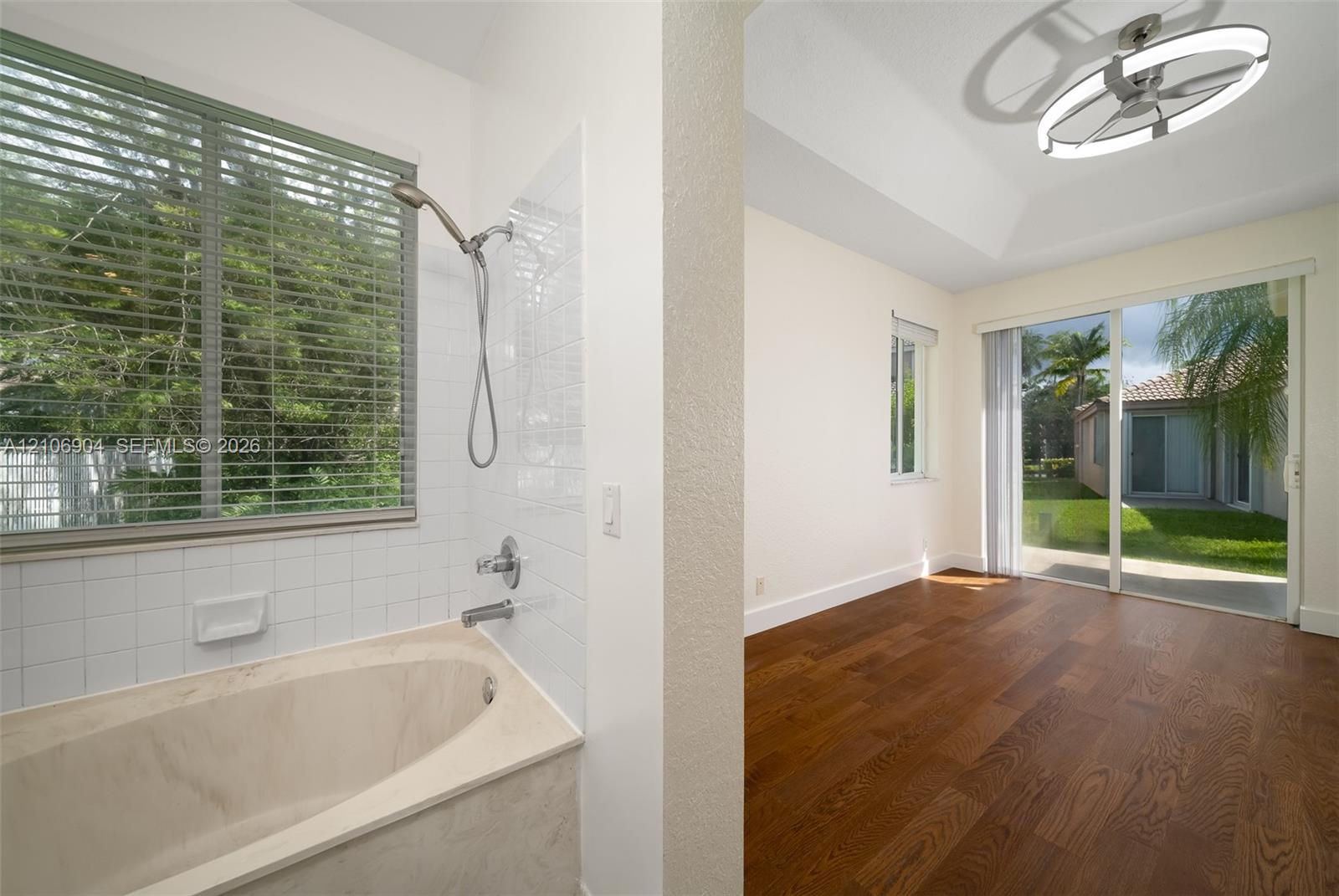 Soaking tub with tile surround, wood-finish flooring, tray ceiling, contemporary circular LED light fixture, and sliding glass doors