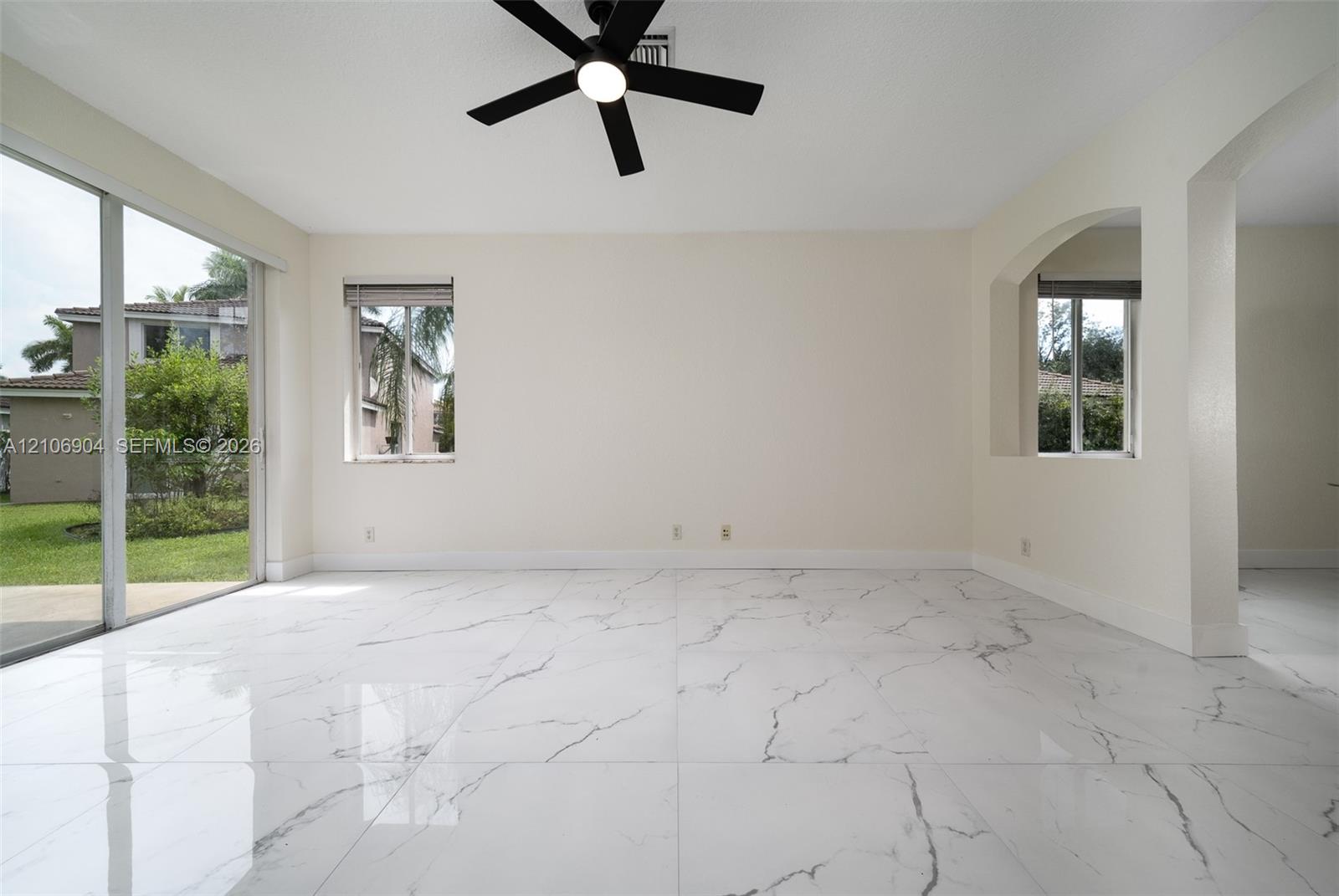 Large-format white tile flooring with grey veining, matte black ceiling fan with integrated lighting, glass sliding doors, and arched architectural wall detail