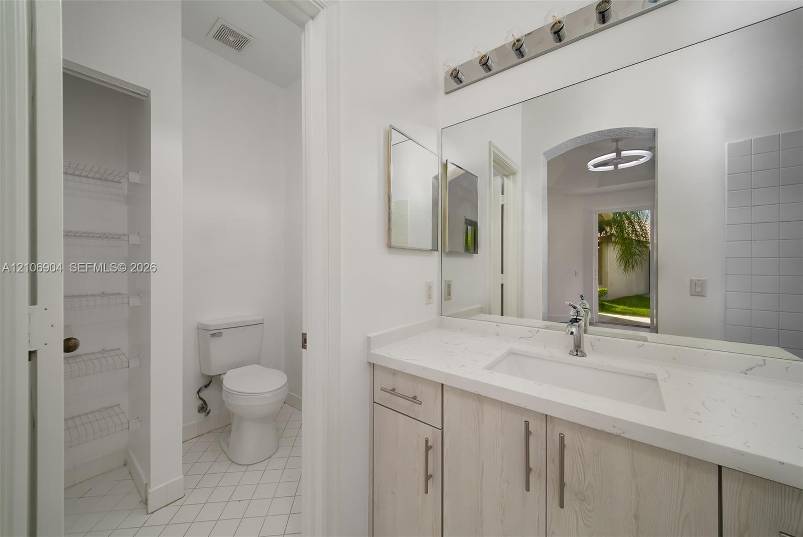 Vanity featuring a light wood-finish cabinet, quartz countertop, and integrated sink, complemented by a large wall-mounted mirror, multi-bulb lighting fixture, and built-in shelving