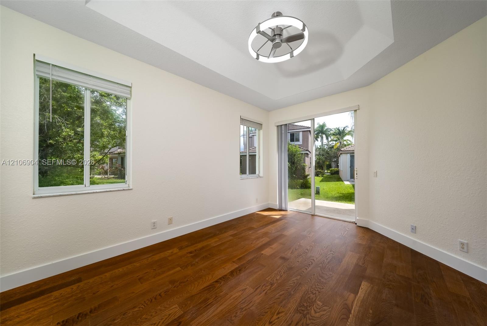 Interior space featuring wood-finish flooring, tray ceiling, integrated circular lighting fixture, two windows with blinds, and sliding glass doors
