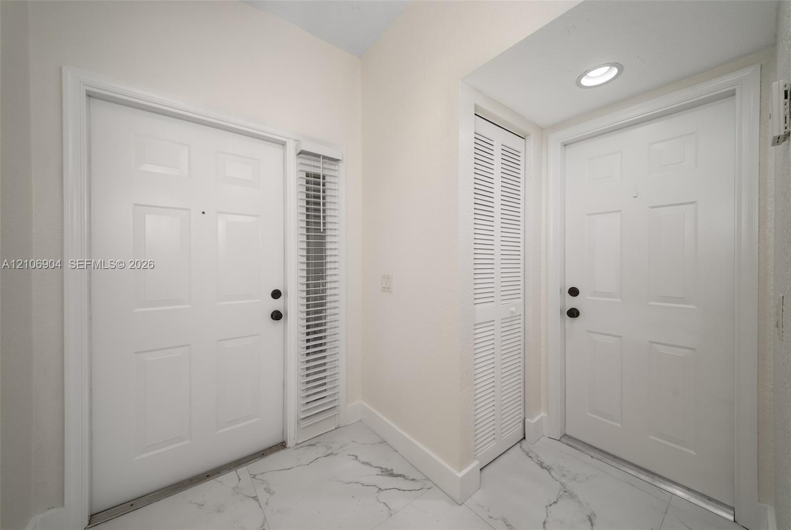Entryway featuring polished marble-style tile flooring, two six-panel doors with matte black hardware, vertical slatted window blinds, and a built-in louvered closet door with recessed ceiling lighting